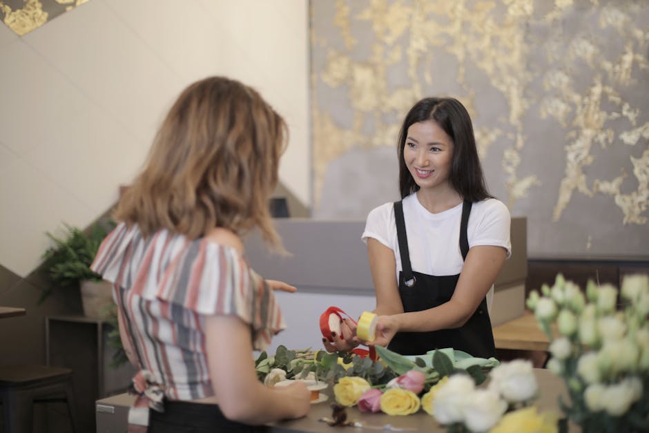 A floral shop employee, wearing a white t-shirt and black apron, is smiling as she hands a floral gift wrapped with a red ribbon and yellow paper to a customer with wavy, shoulder-length light brown h
