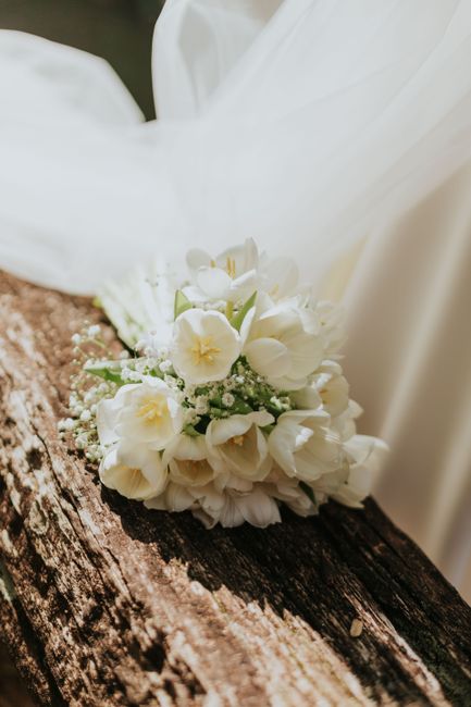 A delicate bouquet of white flowers, including freesia, roses, and baby's breath, rests on a rustic wooden surface. The bouquet features fresh, soft petals and green stems, arranged in a classic style